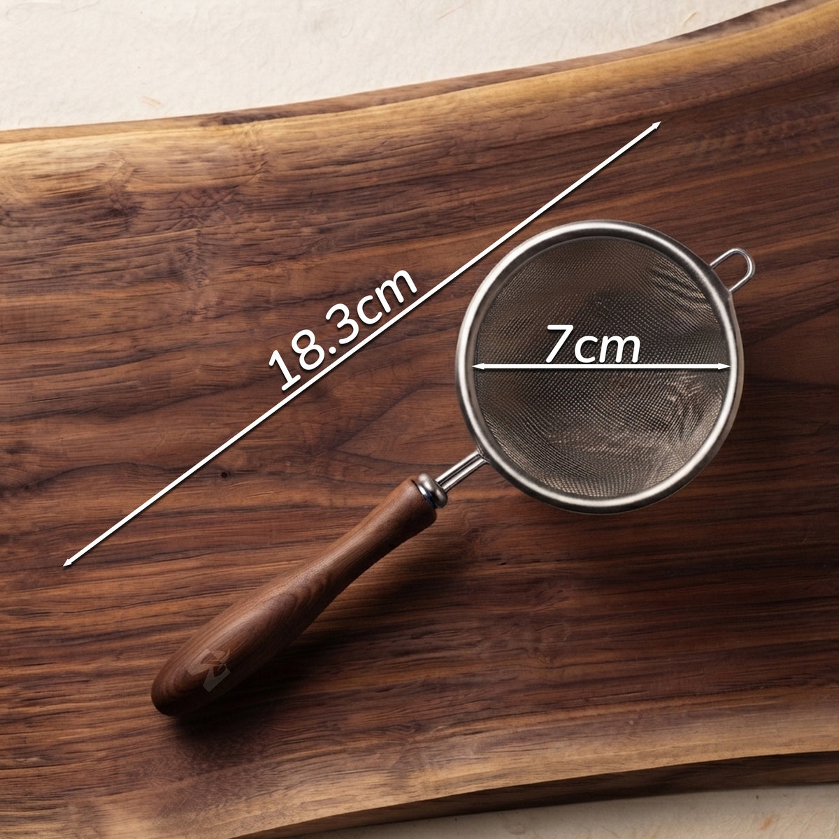 A metal mesh strainer with a dark wooden handle resting on a wooden board, with white measurement lines and text indicating dimensions of 18.3 cm length and 7 cm diameter.